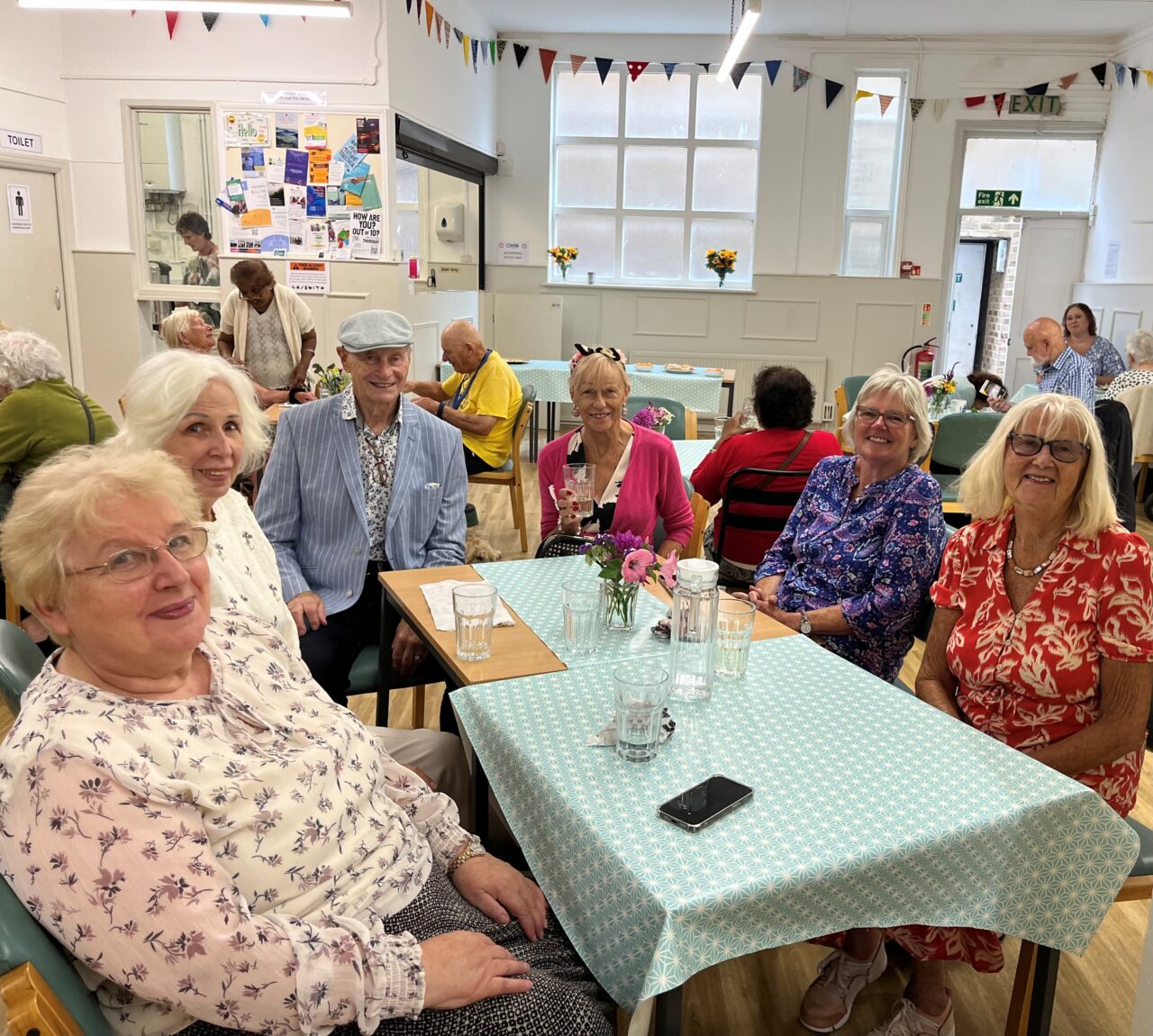 At the Age Friendly Group, several members of the community sitting around a table and smiling at the camera. Other tables are dotted about with other people around them.