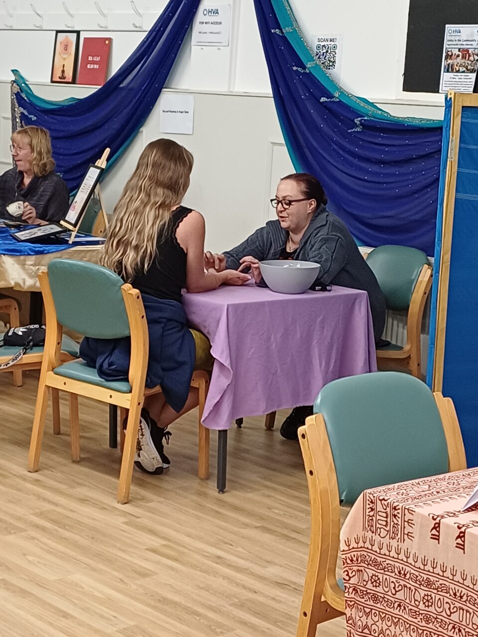 A lady giving another lady a hand massage, sat at a table with a lilac table cloth and a bowl on the table, at a holistic health event