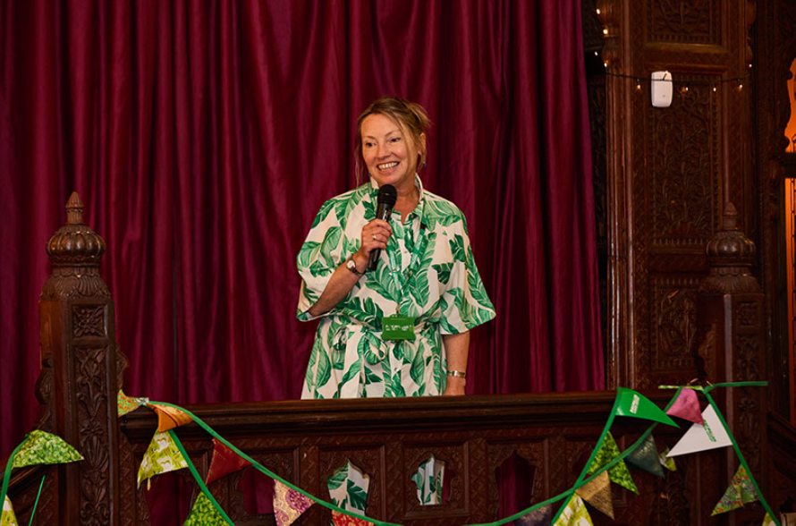 Jane Dodson in a green and white dress, giving a speech, or possible singing. She has a mic in her hand and is smiling. there is bunting draped around.