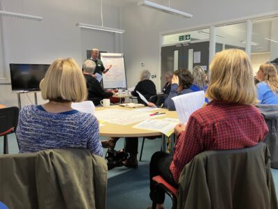 A workshop leader speaks to a room of people with pens and paper on their tables